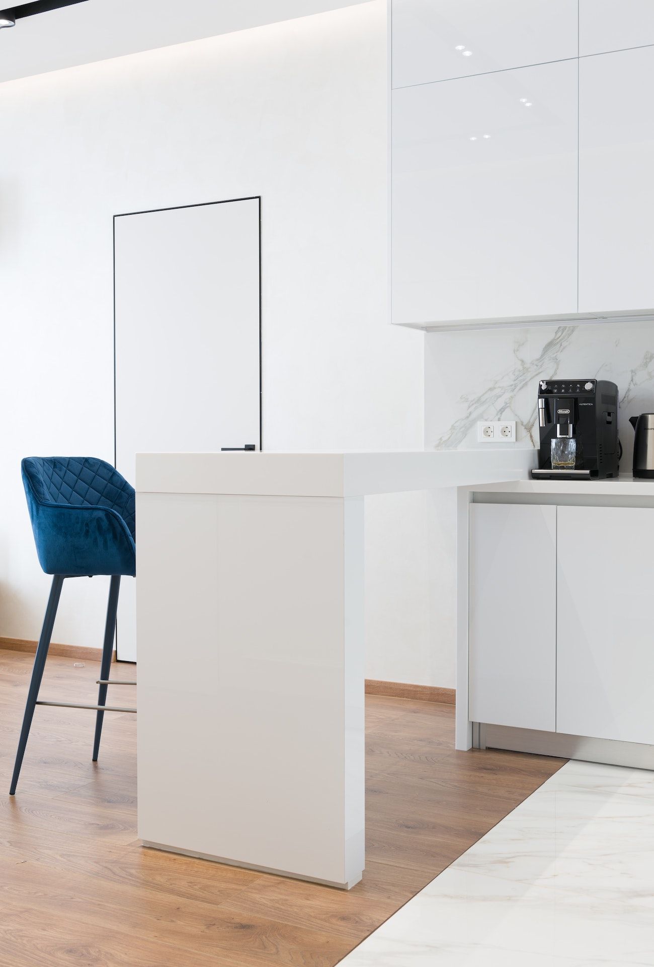 A kitchen with white cabinets , a coffee maker , and a blue chair.