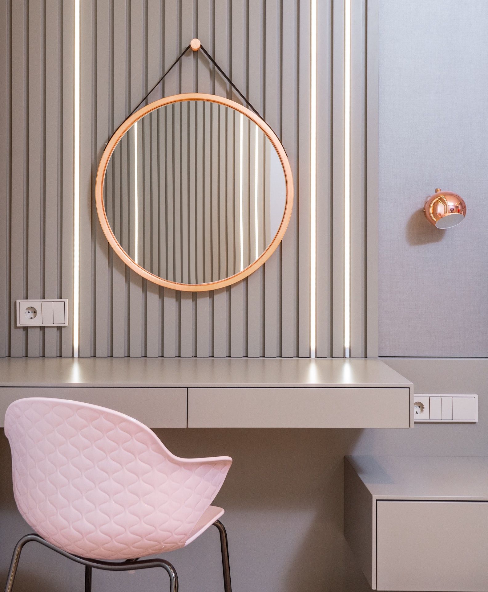 A pink chair is sitting in front of a vanity with a round mirror hanging on the wall.