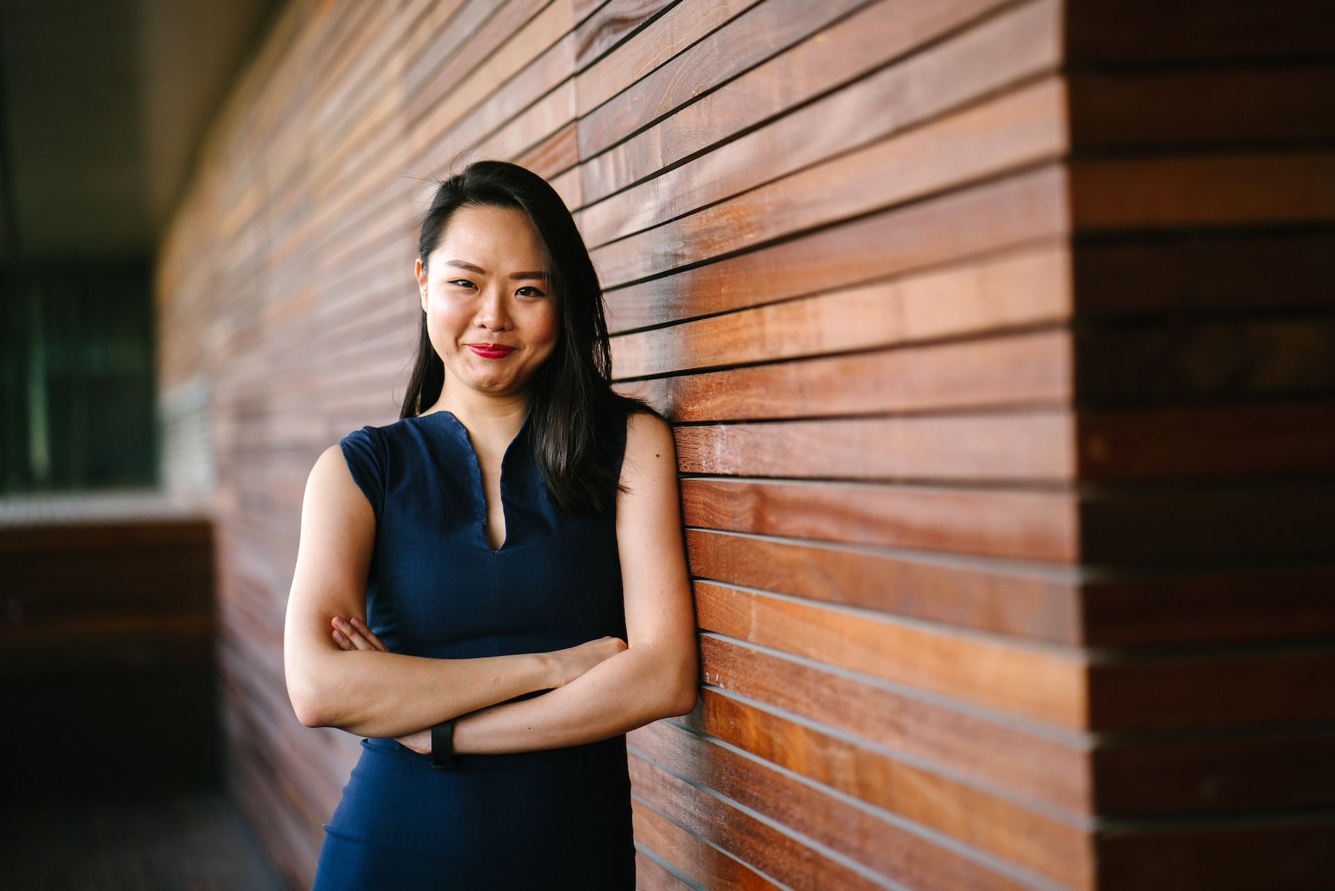 A woman in a blue dress is leaning against a wooden wall with her arms crossed.
