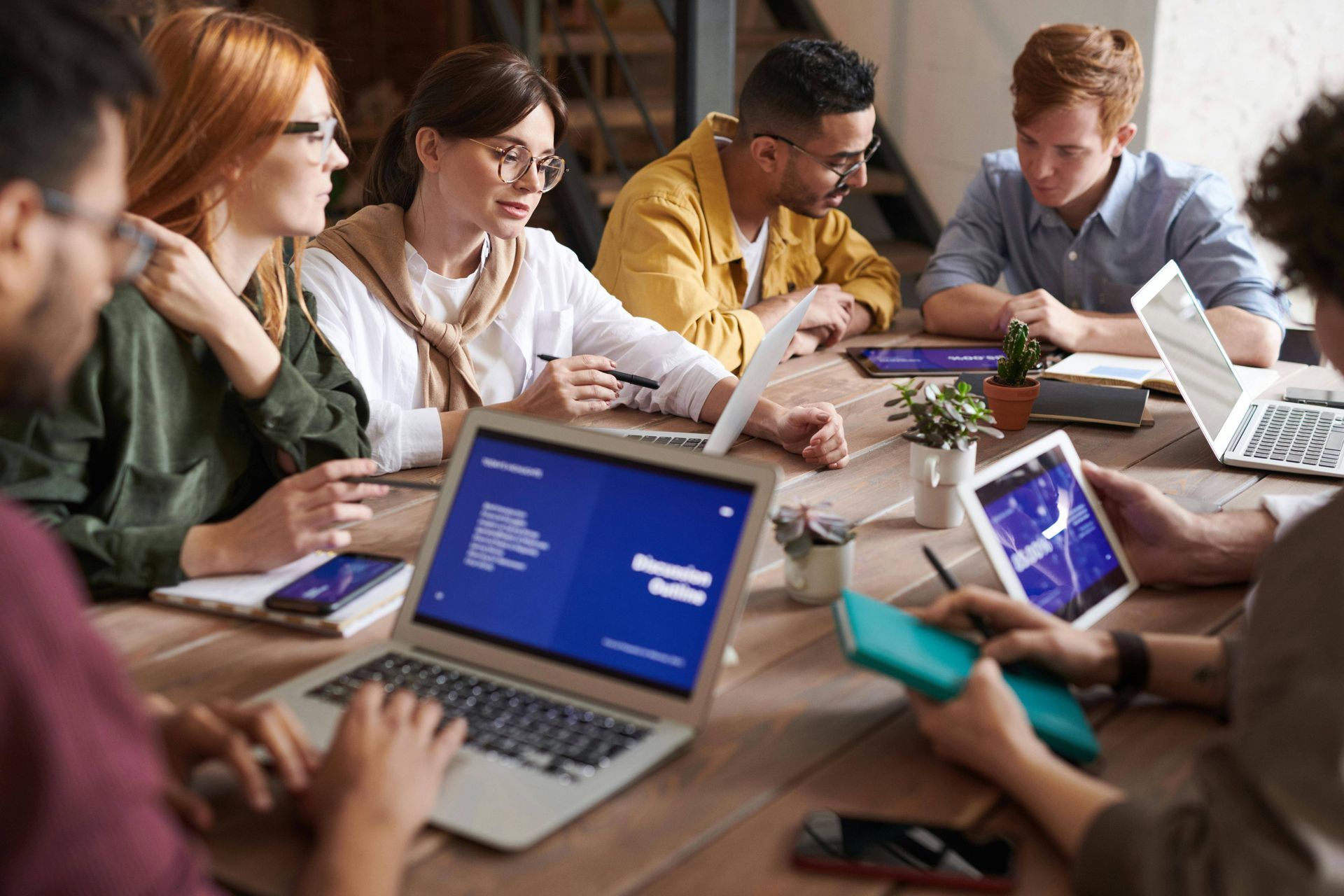 A diverse group of professionals collaborates around a wooden table in an office, using laptops and taking notes.