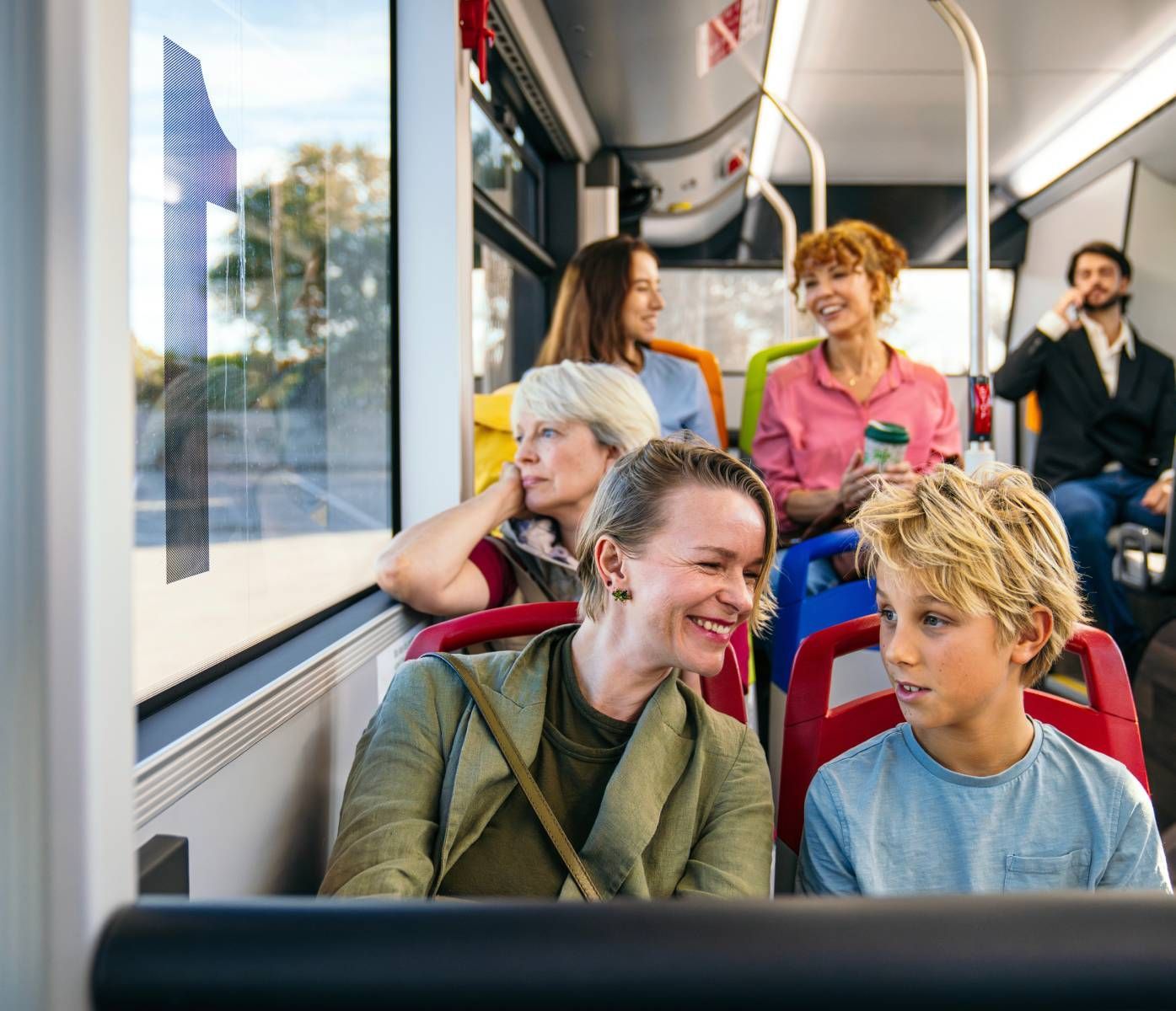 People on a bus, conversing. Woman and child in foreground, others seated nearby. Bright interior, sunny outside.