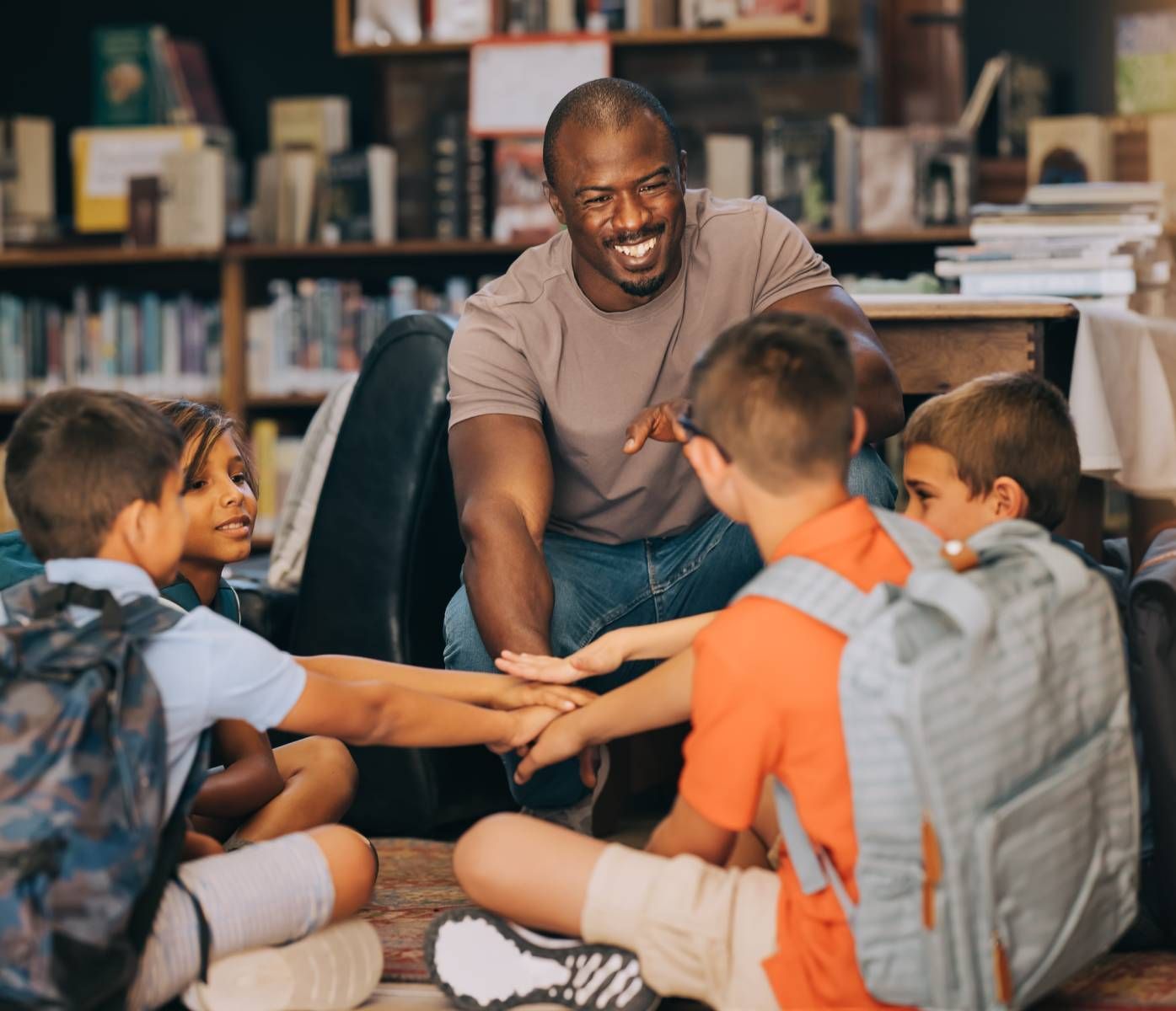 Teacher smiles while high-fiving students. Students in backpacks sit on floor, bookshelved room.