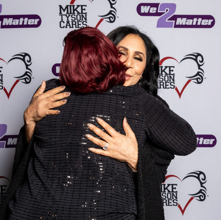 Two women hugging in front of a wall that says mike tyson cares