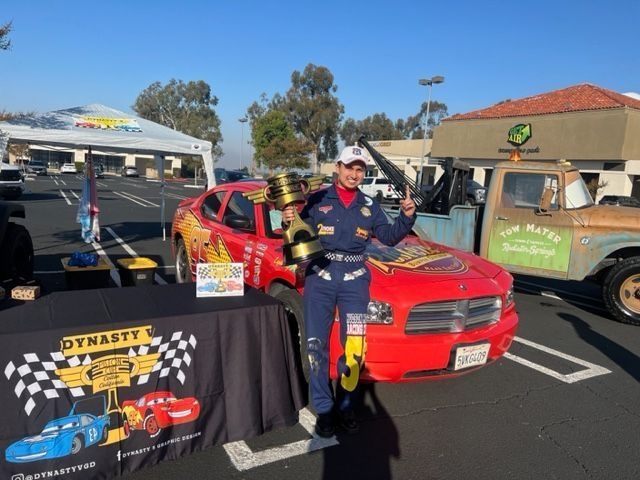 Person in racing suit stands next to a red car, holding a trophy and bottle. Display with "Dynasty" logo.