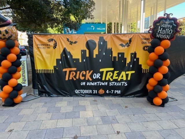 Banner for Halloween "Trick or Treat" on downtown streets. Includes balloon columns, bats, and cityscape silhouette.