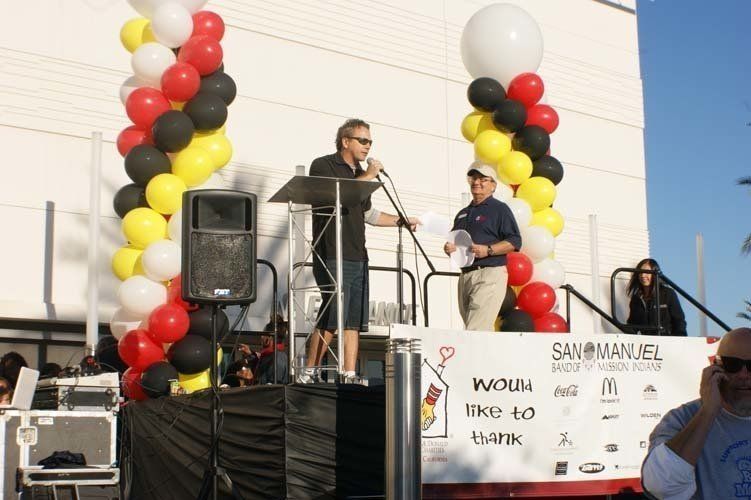 Man speaking at podium on stage, with balloons and logos behind him.