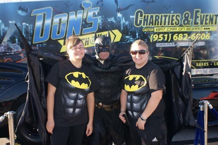 Batman poses with two people wearing Batman shirts in front of a charity event banner.