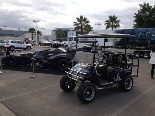 Black golf cart with large tires parked in front of a dark car on a trailer, outside.