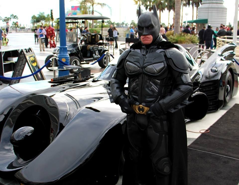 Batman in costume poses next to the Batmobile in an outdoor setting.