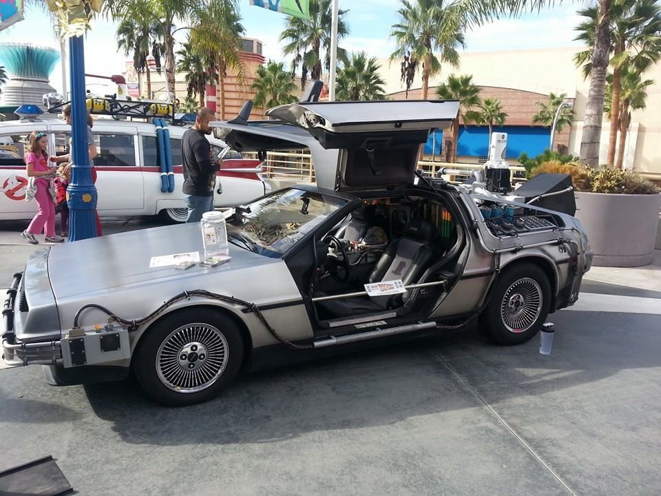 DeLorean time machine with gull-wing doors open, parked outdoors near a Ghostbusters vehicle.
