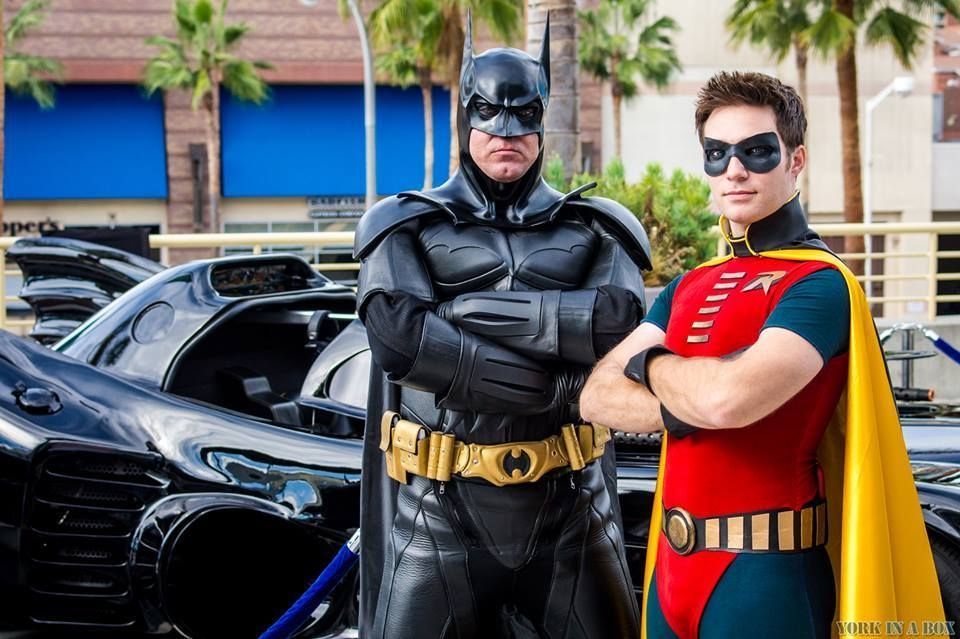 Batman and Robin cosplayers pose by a Batmobile; Batman has crossed arms, Robin stands beside him.