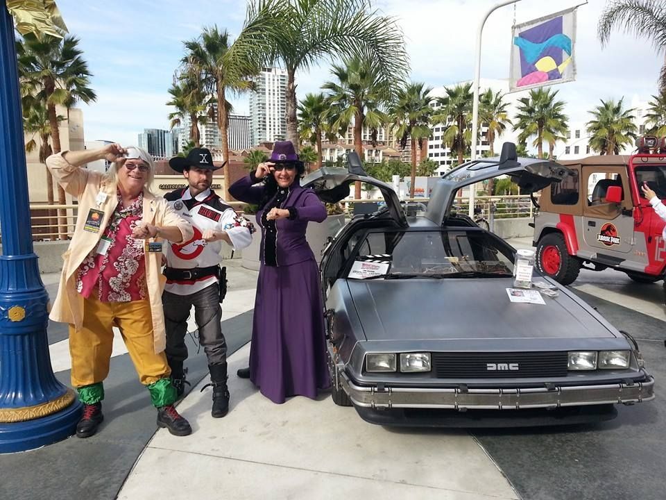 Three people in costumes pose with a DeLorean. One is dressed as Doc Brown, one as a Ghostbuster, and another in purple.