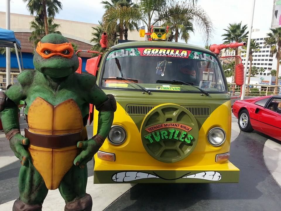 Michelangelo statue posing next to a yellow and green Teenage Mutant Ninja Turtles van. Outdoor setting.