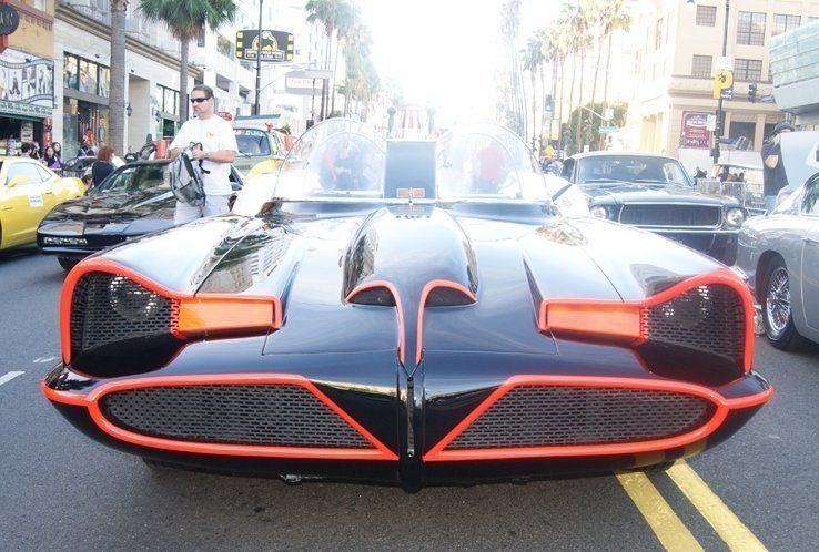 Batmobile on a city street, black with orange trim, surrounded by other cars and people.
