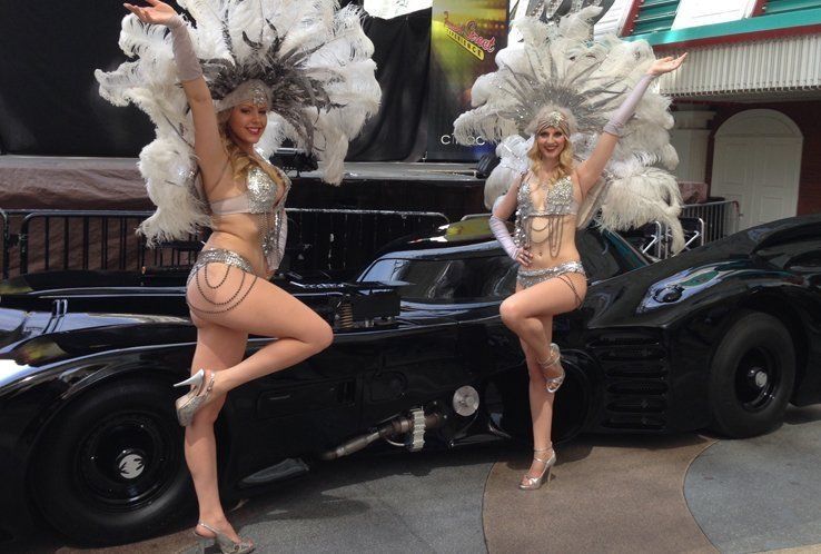 Two showgirls pose by a black Batmobile; both wear silver costumes and headdresses, arms raised.