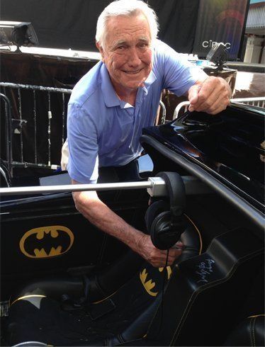 Man smiles, leans into Batmobile, touching seat; Batman logo on car. Outdoors.