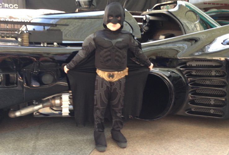 Child in a Batman costume stands in front of the Batmobile, holding his cape open.