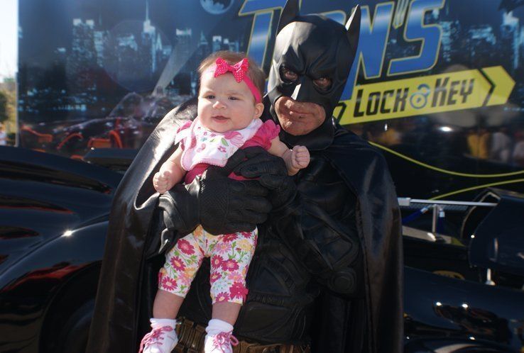Batman in costume holding a smiling baby, next to a car with “Torn's Lock & Key” sign.