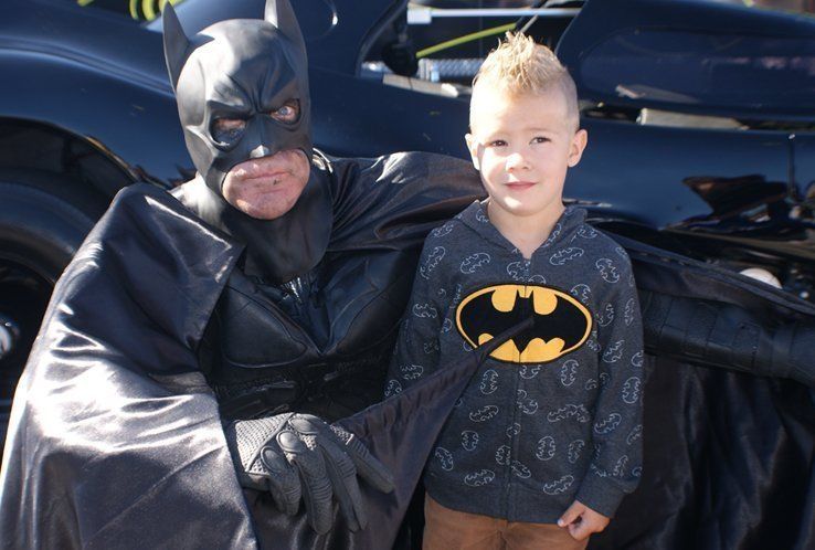 Batman in costume poses with a young boy wearing a Batman hoodie near a Batmobile.
