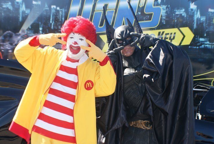 Ronald McDonald and Batman posing together, making peace signs, near a car.