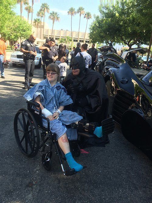 A person in a wheelchair with a leg injury sits with Batman next to a Batmobile; people and palm trees in background.