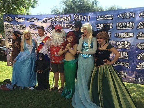 Group of costumed characters pose in front of a banner. Includes Elsa, Ariel, Anna, Batman, and a Santa-like figure.