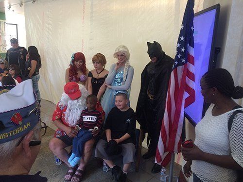 Costumed characters with children and flag at an event, including Santa, Batman, and Elsa.