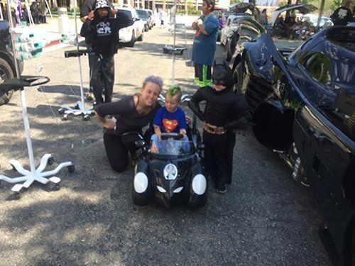 Woman and two children in superhero costumes pose near a Batmobile and toy car; street setting.