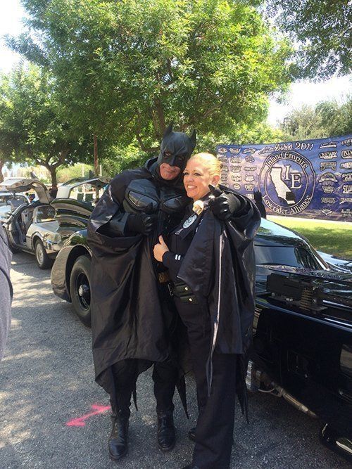 Batman in costume hugs a woman near a Batmobile and other black cars outdoors.