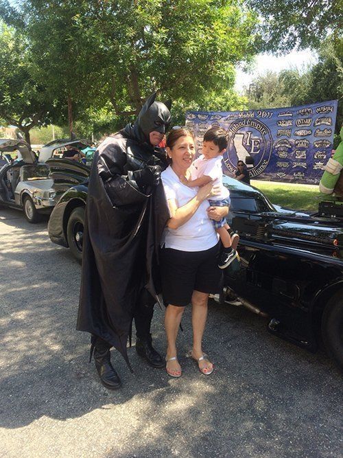 Batman poses with a woman and child next to the Batmobile at an outdoor event.