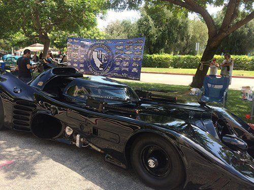 A black Batmobile replica is on display at an outdoor event with a blue banner and people nearby.