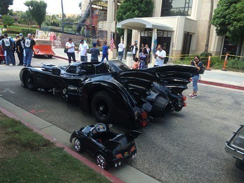 A full-size Batmobile next to a child's toy version on a street, with people nearby and a building in the background.