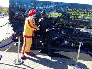 Ronald McDonald shakes hands with Batman in front of a themed vehicle.