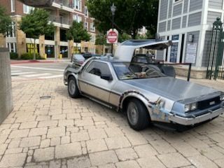 Delorean car with gull-wing doors open, parked on a brick-paved sidewalk near a street corner and buildings.
