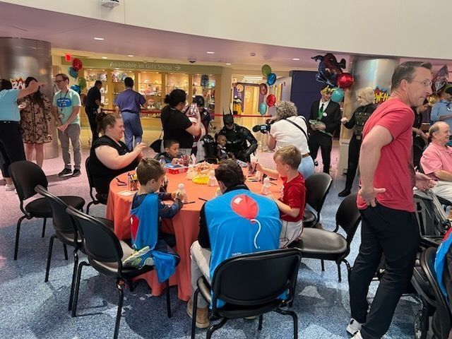 Children in superhero costumes at a table, interacting with people in a large room.