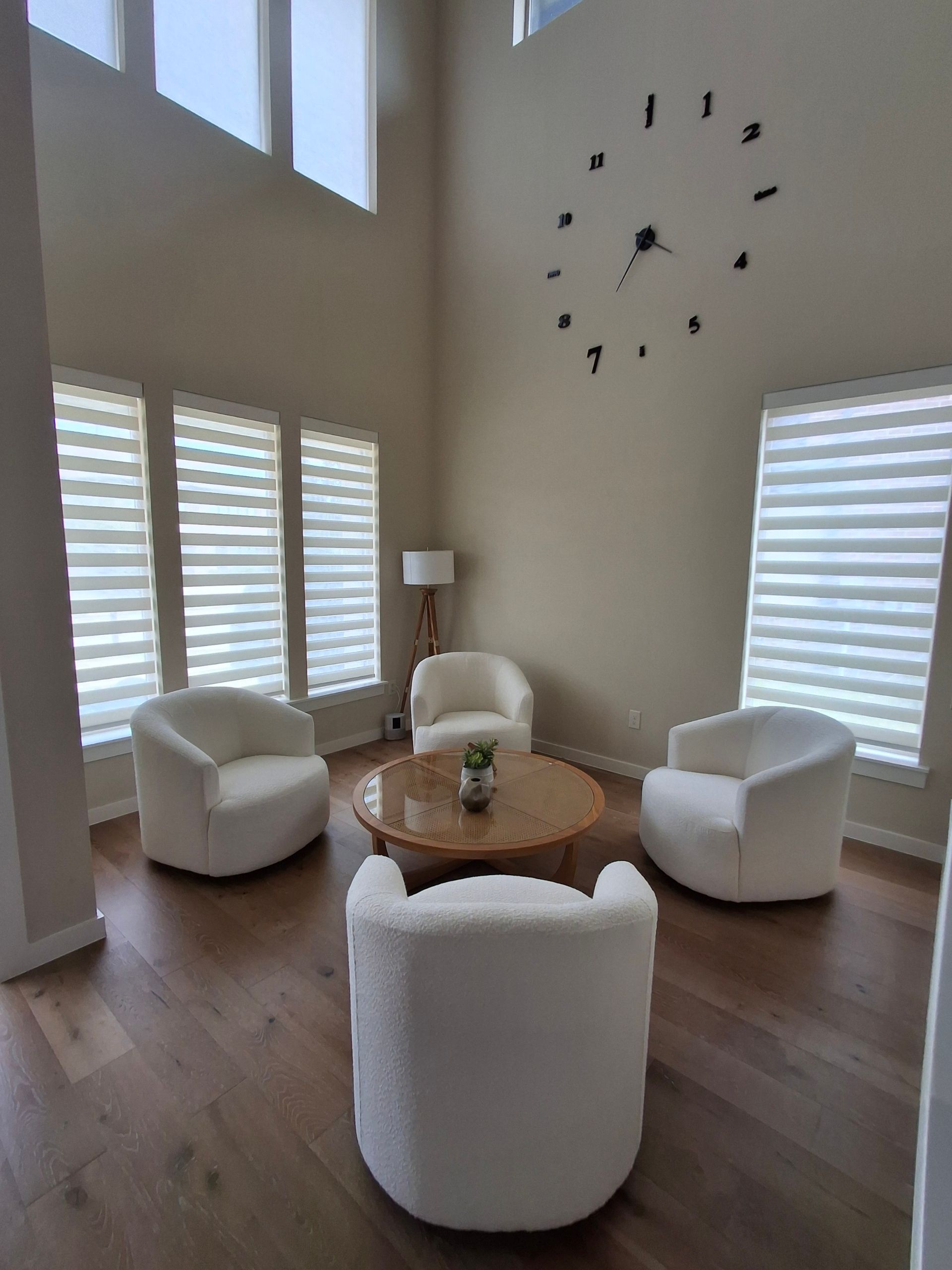 Living room with four white chairs around a wooden table, large clock on the wall, and tall windows.