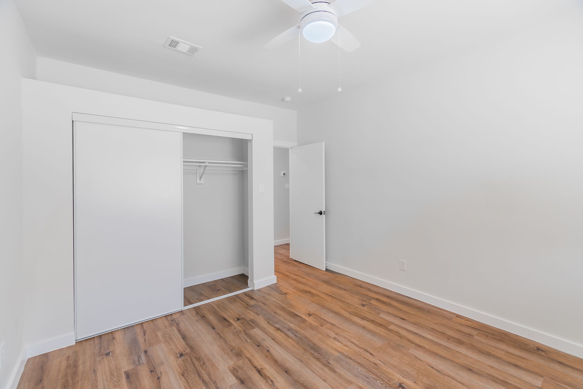 Empty bedroom with white walls, wood floor, closet, and ceiling fan.