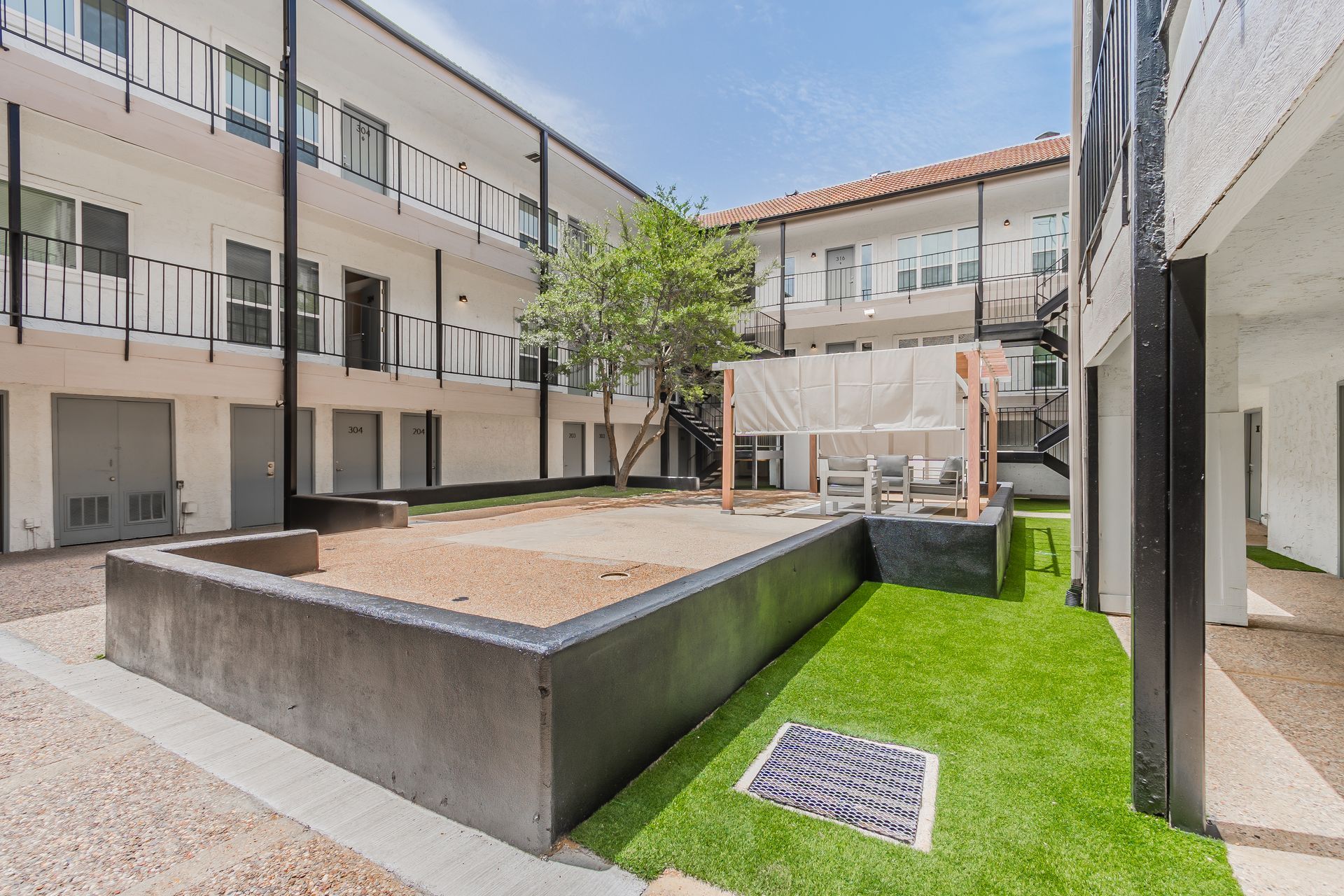 Courtyard of a white apartment building, with a sandbox, fake grass, and a tree.