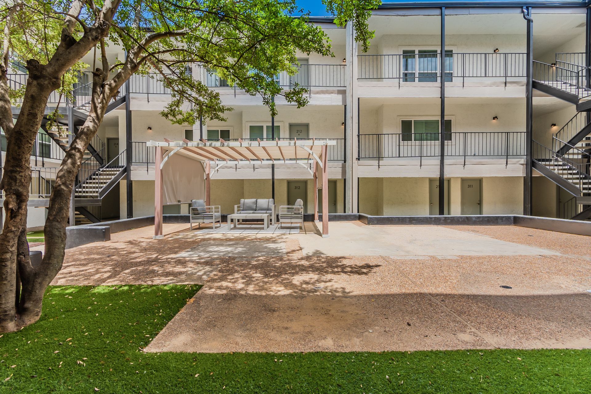 Courtyard of an apartment building with seating under a pergola, gravel ground, and artificial grass.