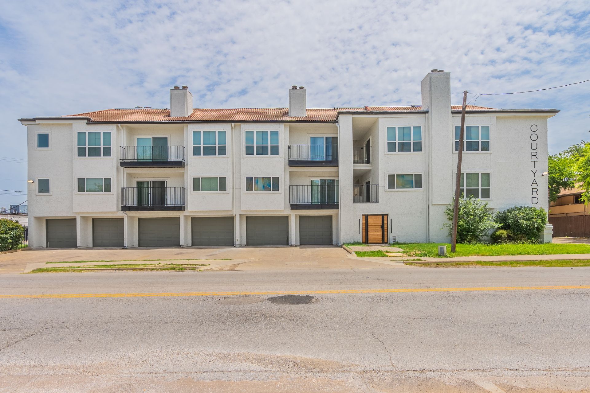 White condo building with garages, balconies, and a terracotta tile roof along a road.