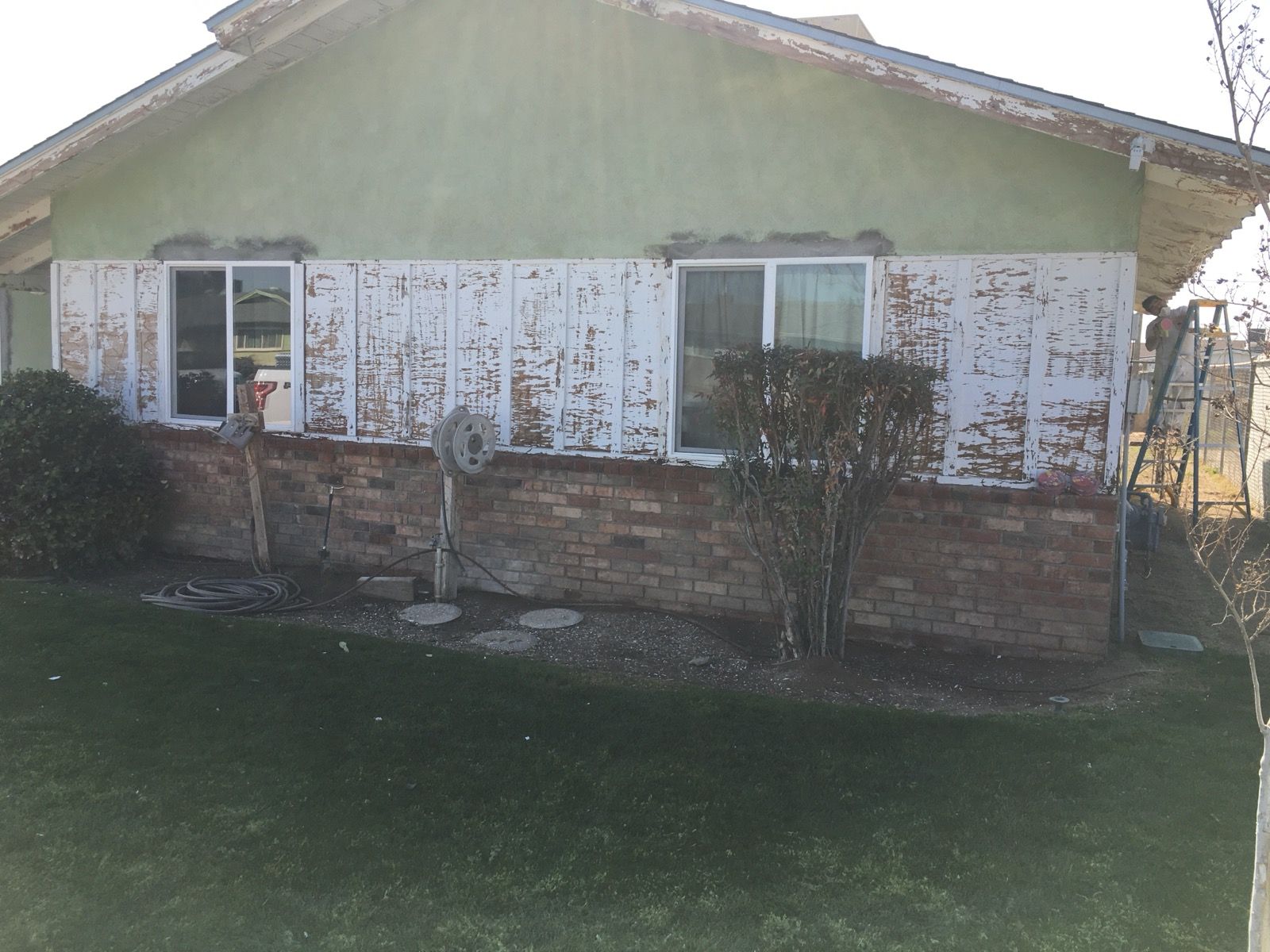 Exterior view of a house with peeling white paint, two windows, and brick base.