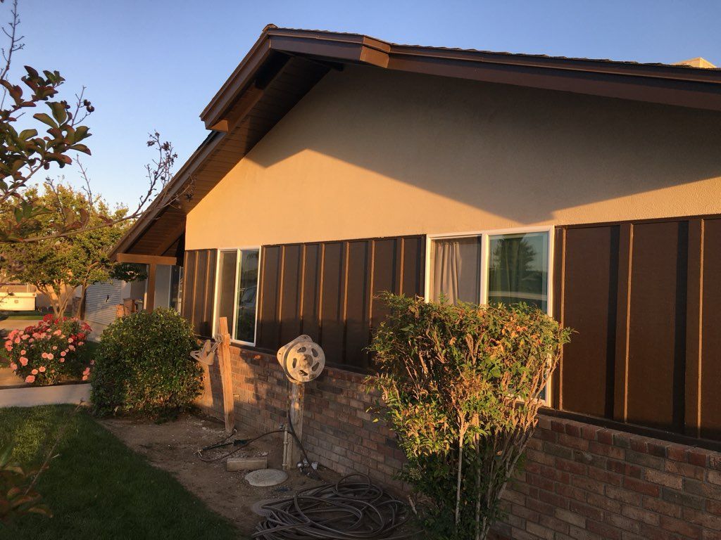 Brown-sided house with windows and tan stucco, bushes, and a brick wall.