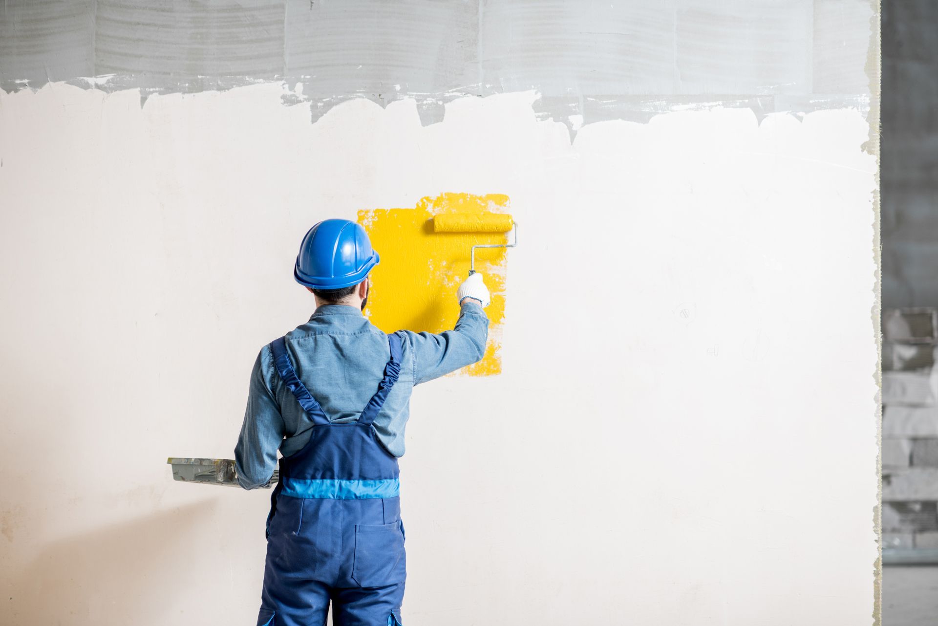Person in blue overalls and helmet painting a yellow square on a white wall with a roller.