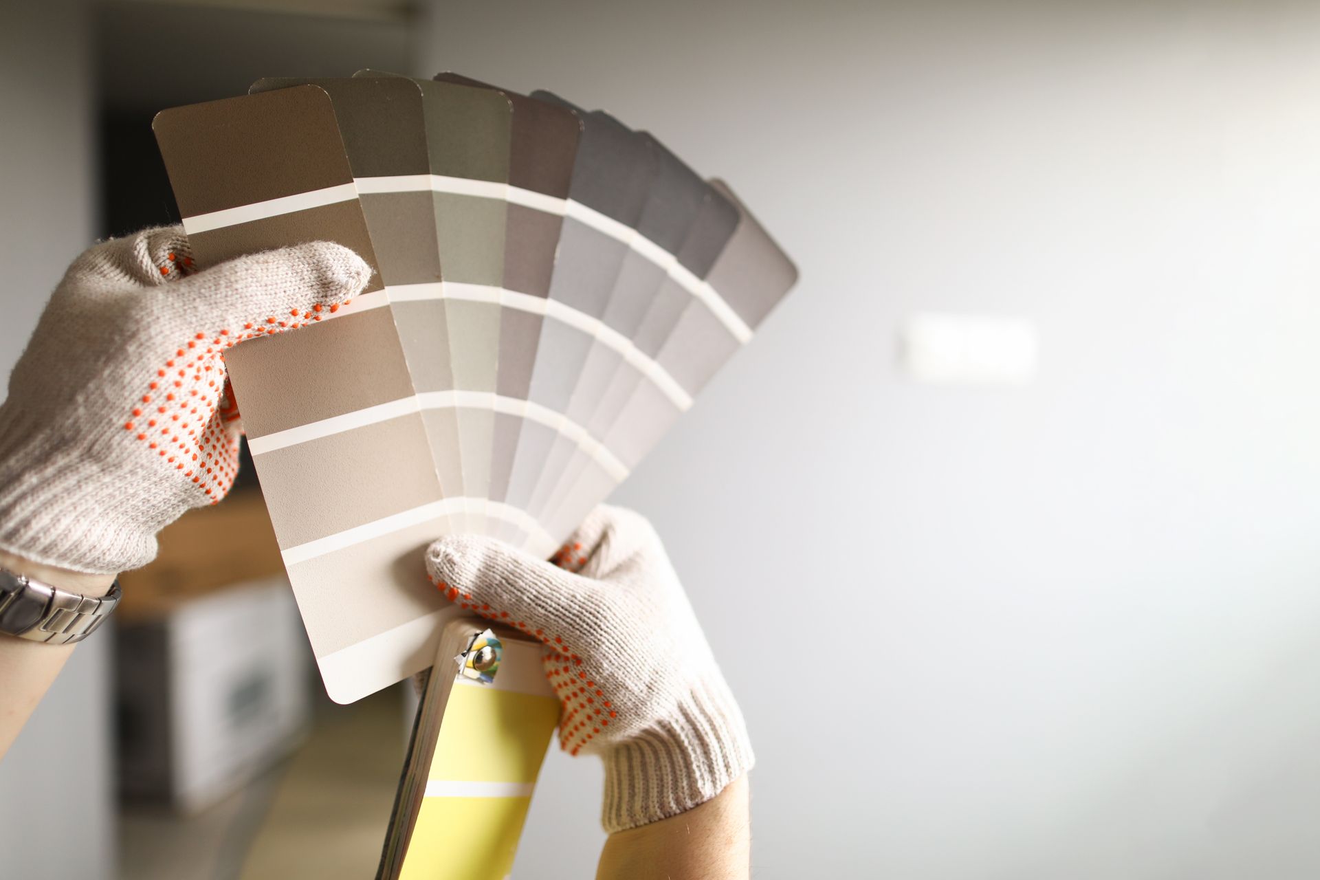 Hands in work gloves holding a color swatch fan of neutral paint samples against a gray wall.