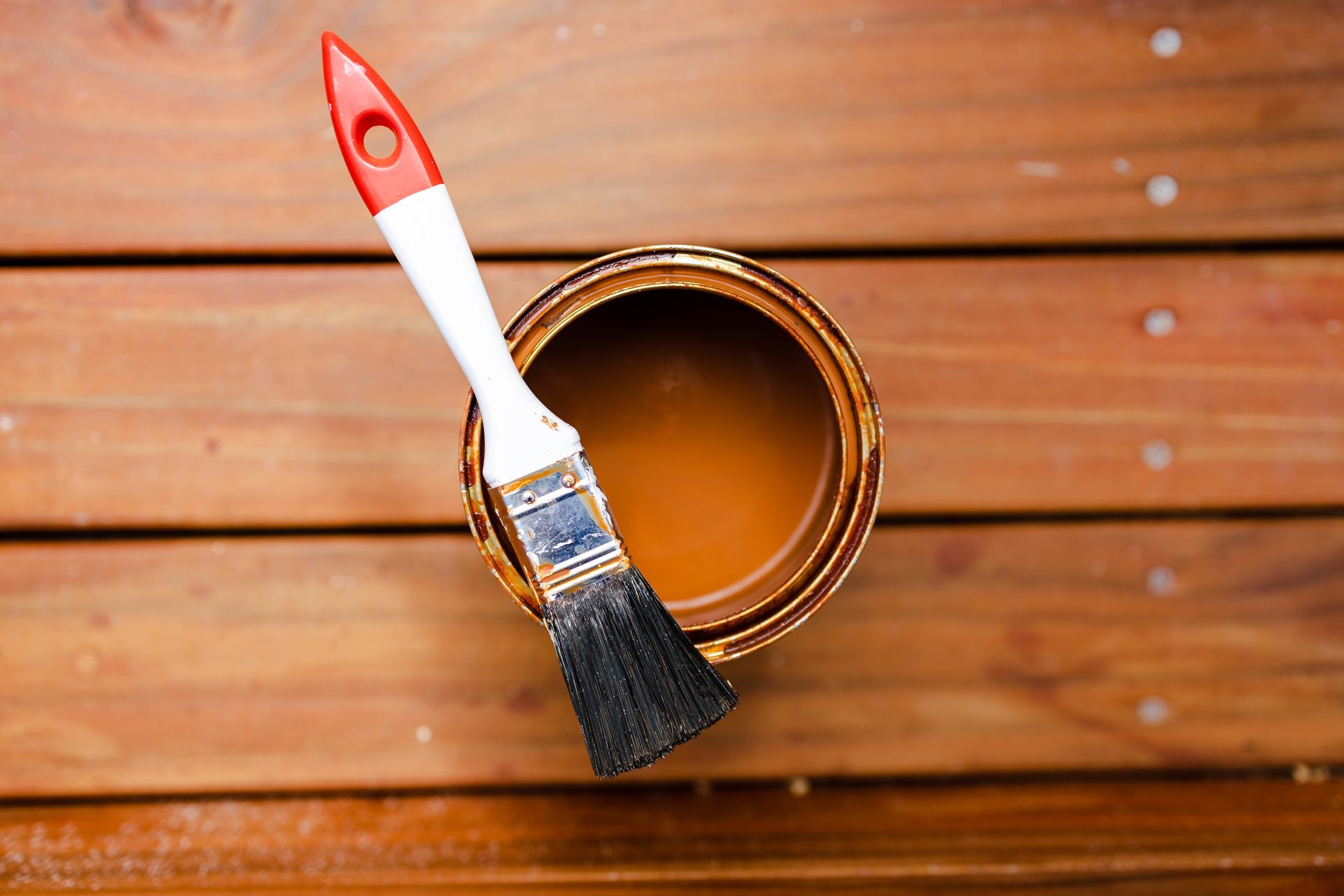 Paint can with brown paint and paintbrush resting inside on wooden deck.