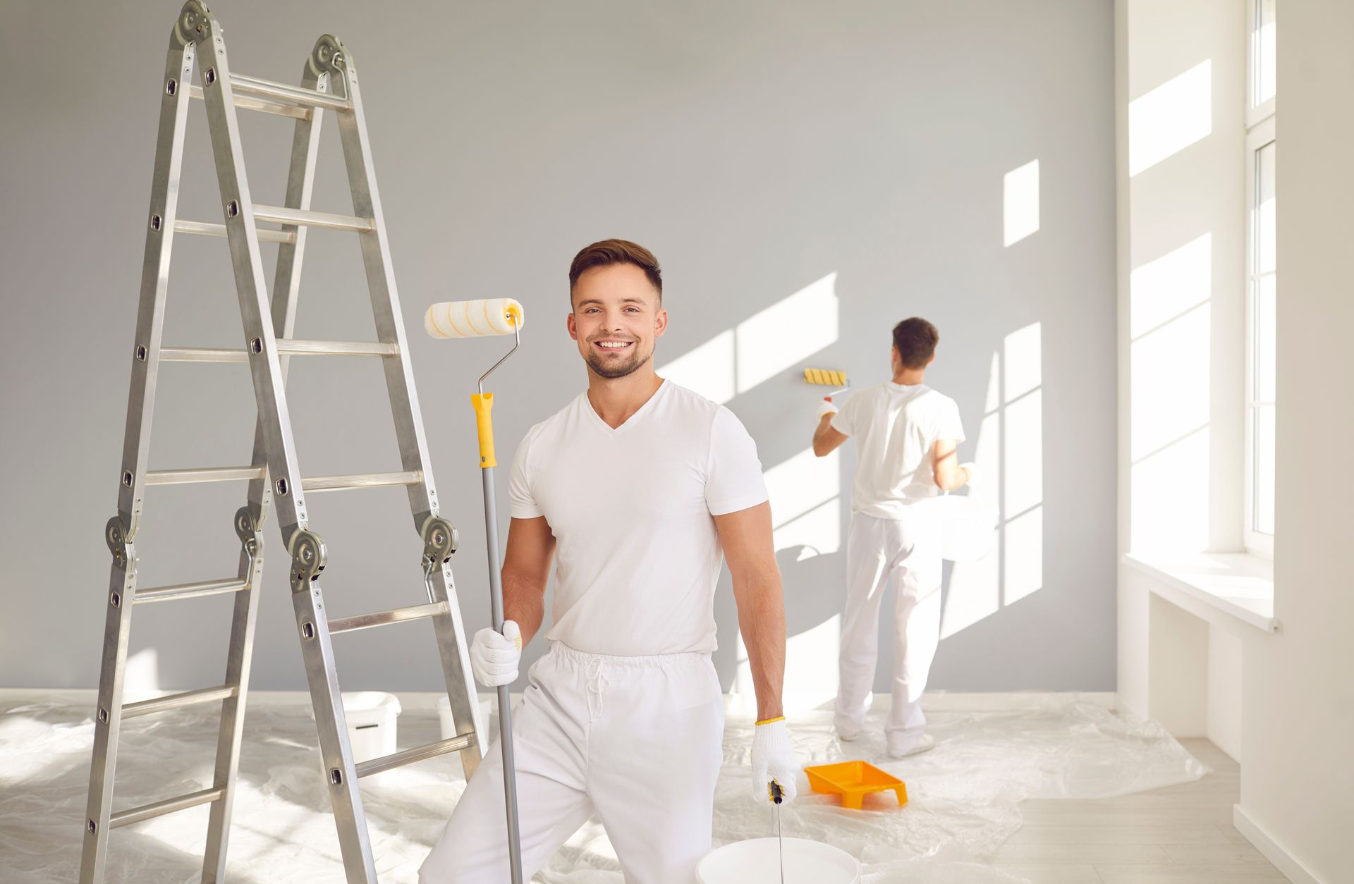 Man in white overalls smiles, holding paint roller.