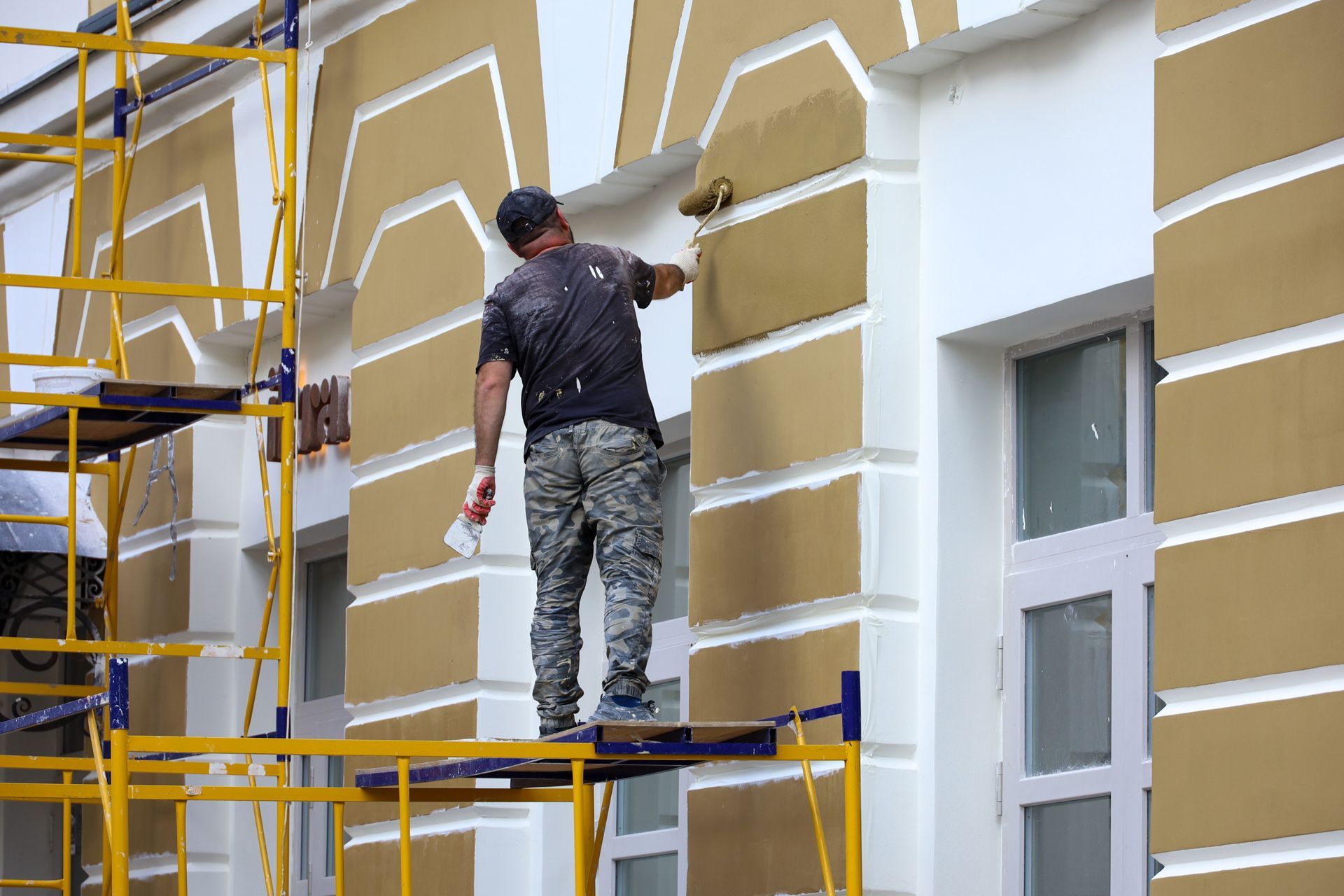 Painter on scaffolding applies yellow paint to building trim.