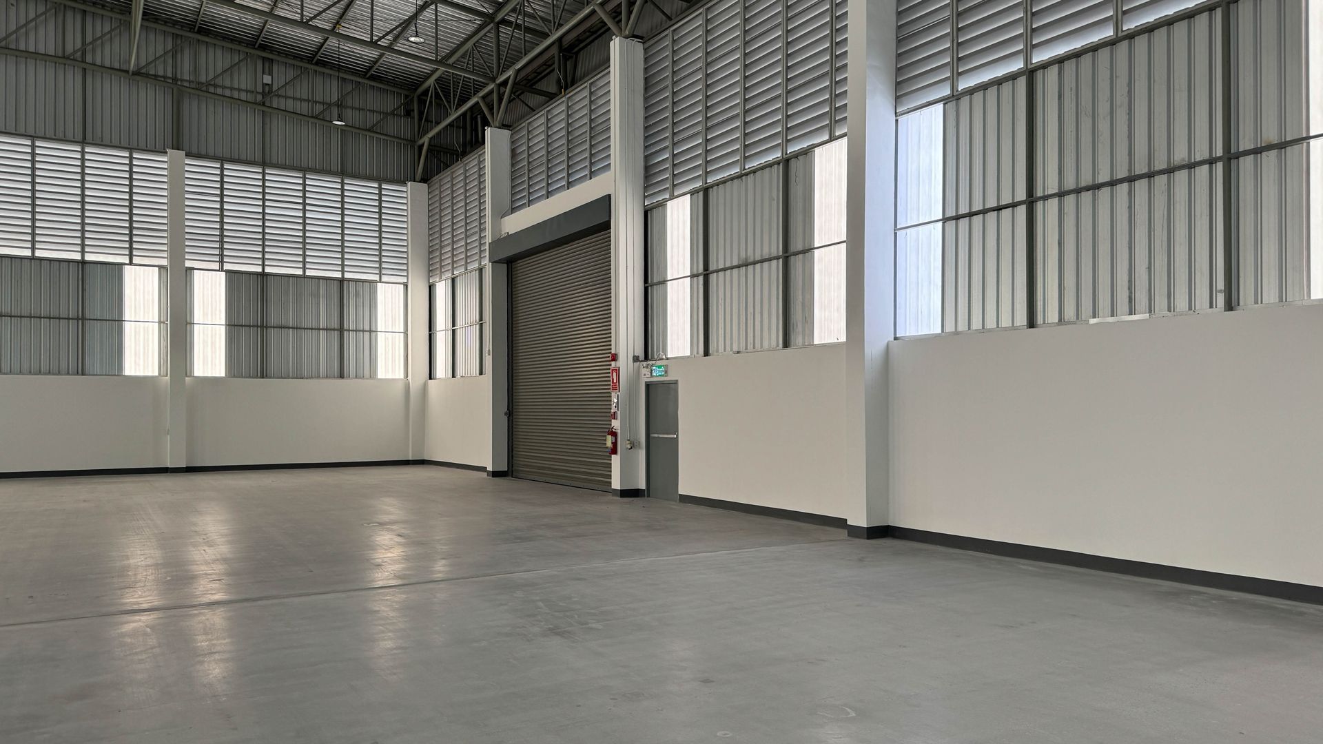 Empty industrial warehouse with grey floor, white walls, metal ceiling, and closed roll-up door.