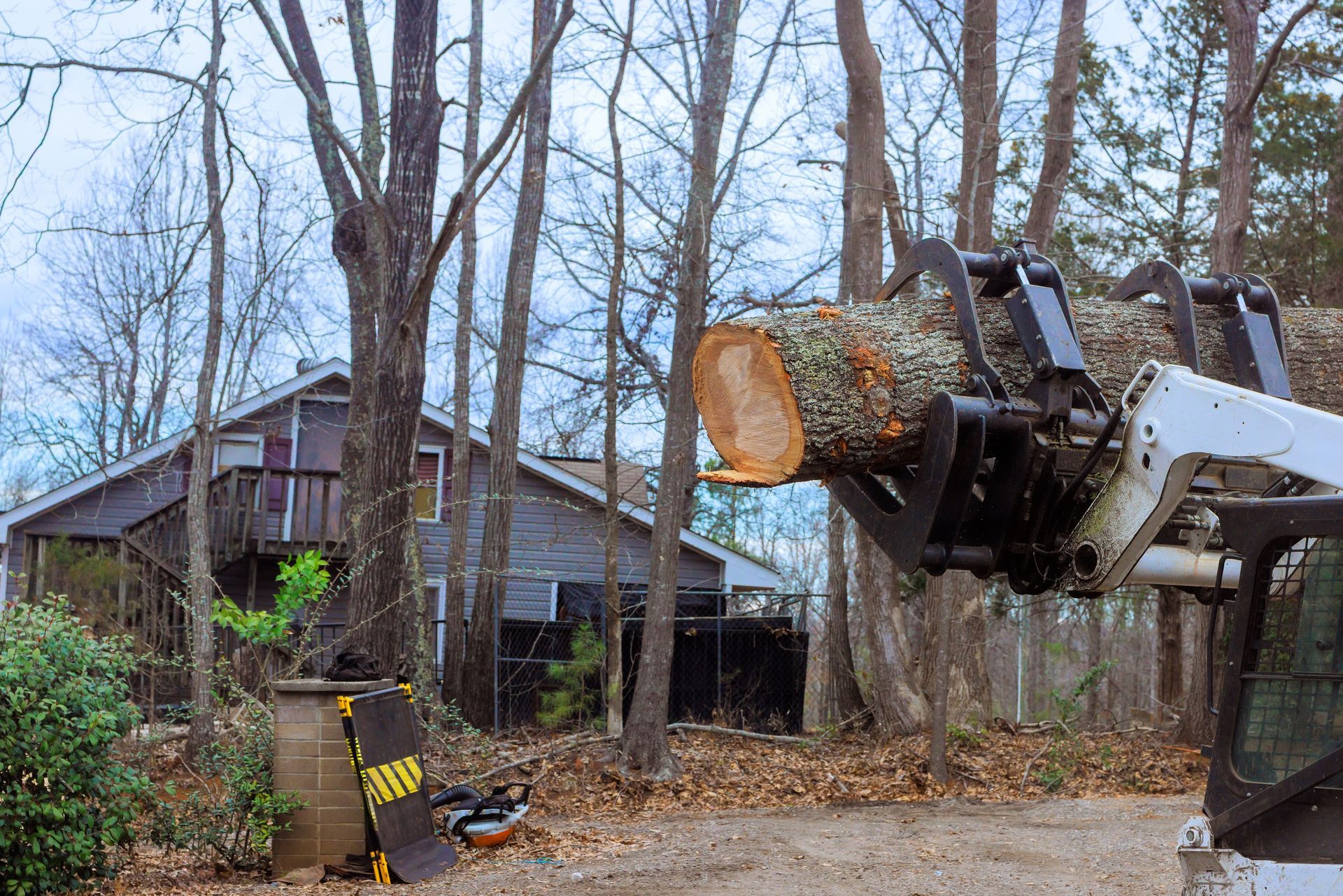 A Bobcat machine lifting a large tree log near a house in a wooded area.