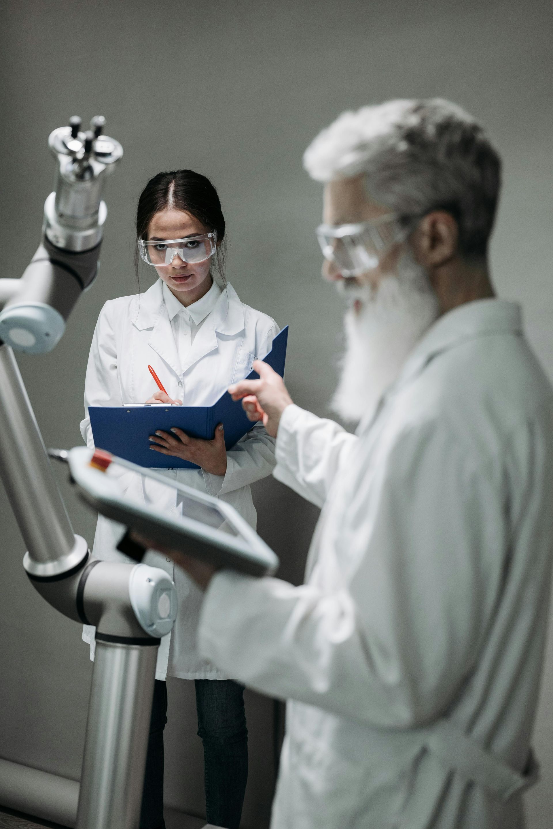 Twee wetenschappers in witte stofjassen en veiligheidsbrillen bedienen een robotarm met een tablet en een klembord.
